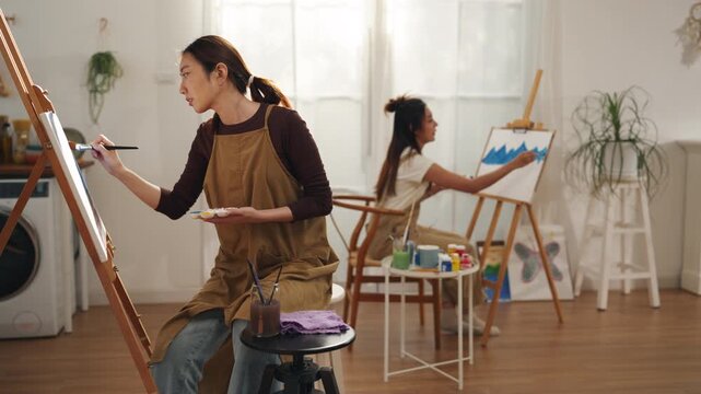 Creative young Asian woman artist painting on canvas with brush in bright home studio. Relaxed female hobbyist enjoying peaceful indoor leisure time, expressing imagination for art.