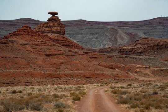 View of the iconic Mexican Hat Rock formation rising above the red desert landscape with a dirt road leading towards the sandstone cliffs in Oljato-Monument Valley, Utah, United States.