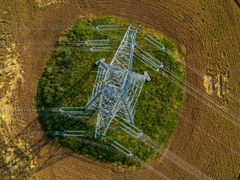 Aerial view of a high-voltage power transmission tower standing in a circular patch of green grass amidst a harvested agricultural field in Stakliskes, Kaunas County, Lithuania.