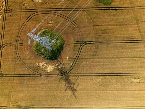 Aerial view of a high-voltage electricity pylon in a green patch within a golden wheat field, casting a long shadow across the agricultural landscape in Stakliskes, Kaunas County, Lithuania.