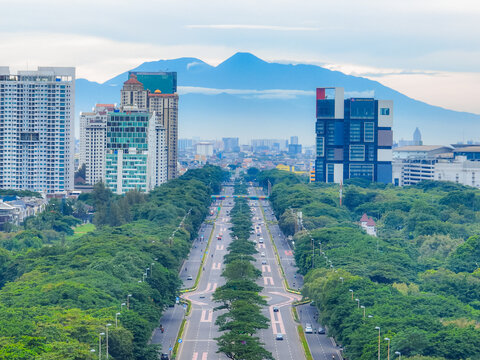 Aerial view of a wide boulevard lined with lush green trees and modern high-rise buildings, with a distant mountain range in the background in Kemayoran, Jakarta, Indonesia.