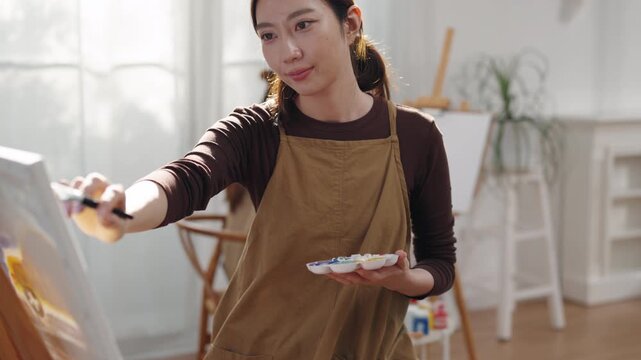 Creative young Asian woman artist painting on canvas with brush in bright home studio. Relaxed female hobbyist enjoying peaceful indoor leisure time, expressing imagination for art.