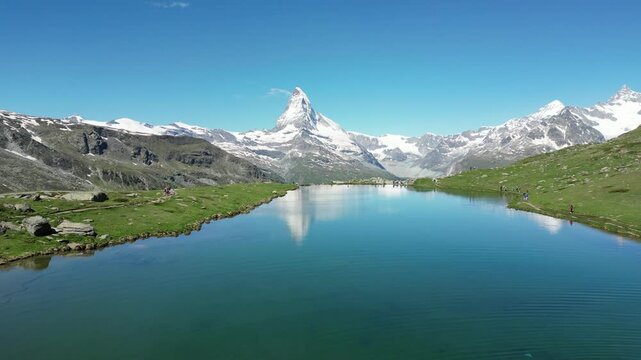 Aerial view of the Matterhorn reflected in the clear waters of Stellisee surrounded by lush green alpine meadows and snow-capped peaks in Zermatt, Valais, Switzerland.