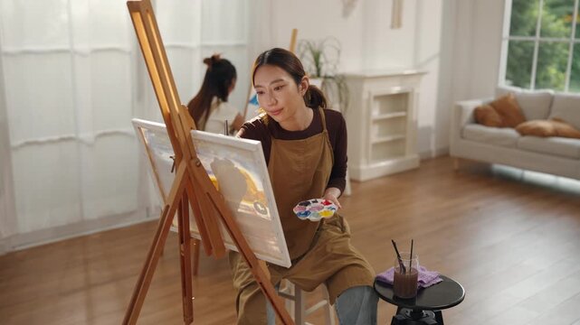 Creative young Asian woman artist painting on canvas with brush in bright home studio. Relaxed female hobbyist enjoying peaceful indoor leisure time, expressing imagination for art.