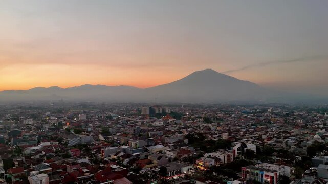 Aerial view of Gunung Arjuno towering over the urban landscape of Malang during a golden sunset, Malang, Jawa Timur, Indonesia.