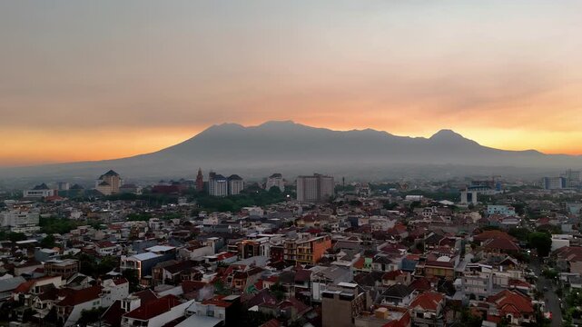 Aerial view of the city of Malang with the silhouettes of Gunung Butak, Gunung Kawi, and Gunung Panderman as one called Barisan Putri Tidur under a golden sunset sky in Malang, East Java, Indonesia.