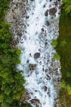 Aerial view of the glacial river in Faueldalen with rushing white water, scattered dark rocks, and lush green forest banks in Jovik, Troms, Norway.
