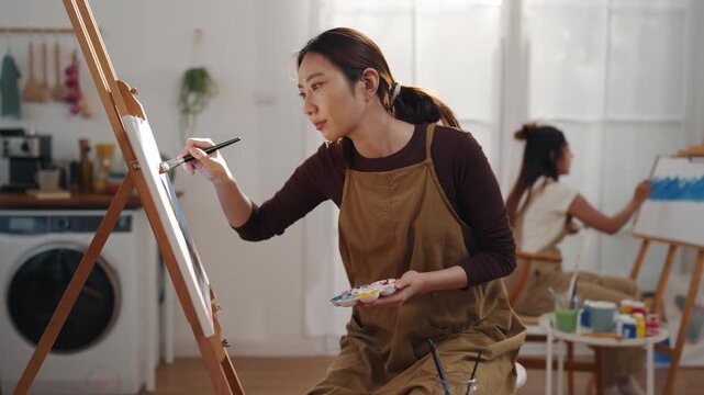 Creative young Asian woman artist painting on canvas with brush in bright home studio. Relaxed female hobbyist enjoying peaceful indoor leisure time, expressing imagination for art.