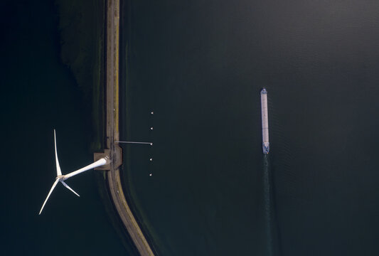 Aerial view of a wind turbine standing on a narrow causeway next to a cargo ship navigating the dark waters of the Krammer locks in Bruinisse, Zeeland, Netherlands.