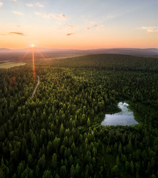 Aerial view of Neitokainen lake shaped like Finland surrounded by dense coniferous forest and a winding dirt road under a golden sunset sky in Kittila, Lapland, Finland.