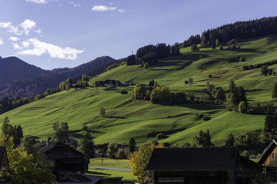 View of lush green mountain slopes dotted with traditional wooden houses, pine forests, and rolling hills under a clear blue sky in Italy.