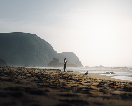 Faro District, Portugal - 20 September 2024: View of a woman standing on a sandy beach near the ocean with dramatic cliffs in the background during golden hour.