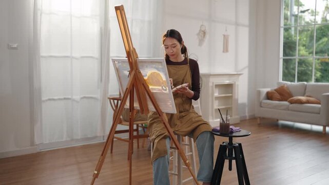 Creative young Asian woman artist painting on canvas with brush in bright home studio. Relaxed female hobbyist enjoying peaceful indoor leisure time, expressing imagination for art.