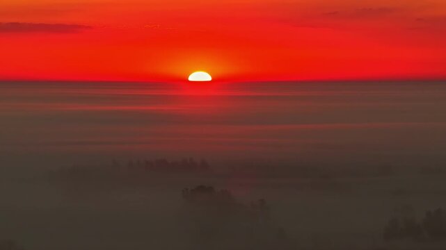 Aerial view of a vibrant red sunset glowing above a thick layer of morning fog covering the rural landscape of LATVIA.