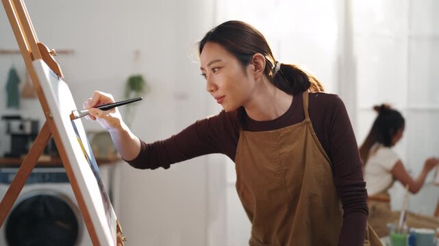 Creative young Asian woman artist painting on canvas with brush in bright home studio. Relaxed female hobbyist enjoying peaceful indoor leisure time, expressing imagination for art.