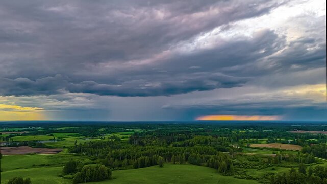 Aerial view of lush green fields and forests under a dramatic, dark, stormy sky with a sliver of golden light on the horizon, Latvia.