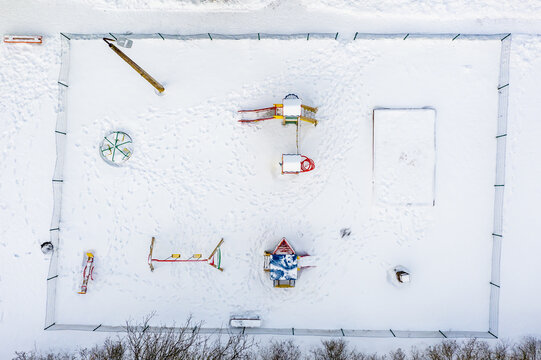Aerial view of a snow-covered playground featuring a slide, a merry-go-round, a swing set, and a sandbox, surrounded by a fence in Latvia.