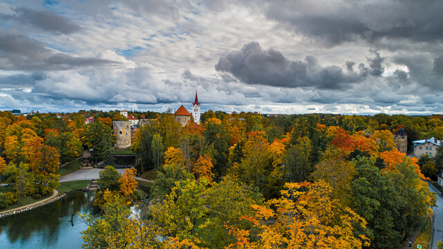 Aerial view of Cesis Castle surrounded by vibrant autumn foliage, historic stone towers, and a park pond under a dramatic cloudy sky in Cesis, Cesis Municipality, Latvia.