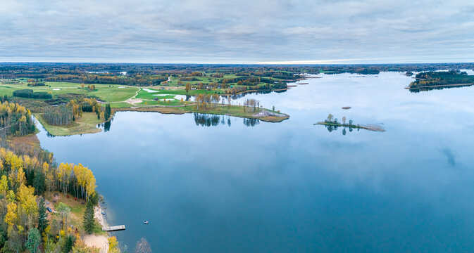 Aerial view of the serene lake in Cesis surrounded by autumn forests, green meadows, and calm waters under a cloudy sky in Cesis, Cesis Municipality, Latvia.