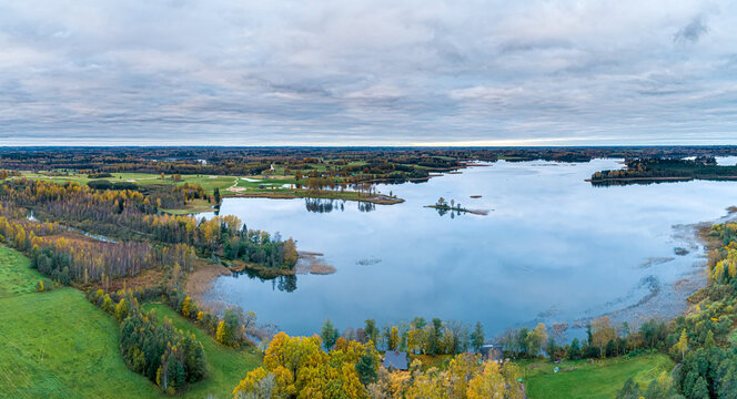 Aerial view of the autumn landscape surrounding the lake in Cesis, featuring vibrant foliage, calm water, and green meadows under a cloudy sky in Cesis, Cesis Municipality, Latvia.