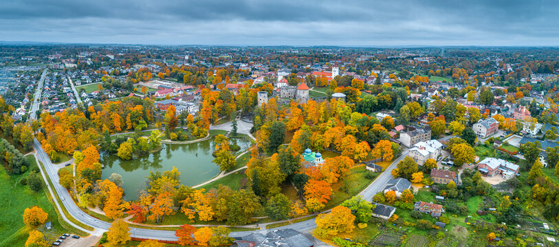 Aerial view of Cesis Castle park surrounded by vibrant autumn foliage, historic stone towers, and urban rooftops under a cloudy sky in Cesis, Cesis Municipality, Latvia.