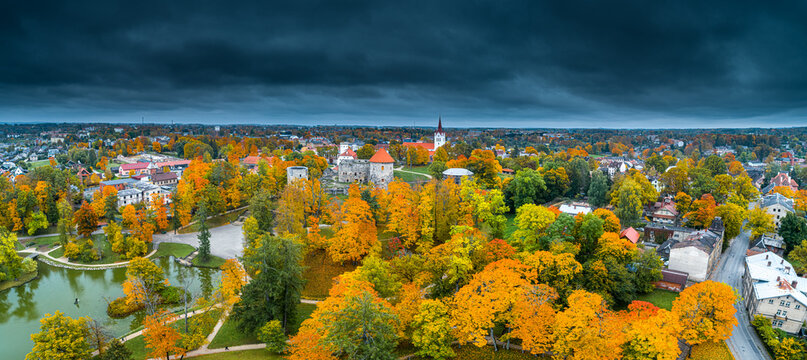 Aerial view of Cesis Castle ruins surrounded by vibrant autumn foliage, historic stone walls, and lush parkland under a dramatic overcast sky in Cesis, Cesis Municipality, Latvia.