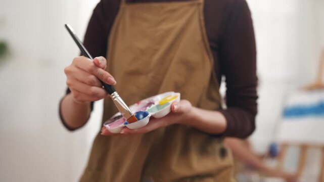 Creative young Asian woman artist painting on canvas with brush in bright home studio. Relaxed female hobbyist enjoying peaceful indoor leisure time, expressing imagination for art.