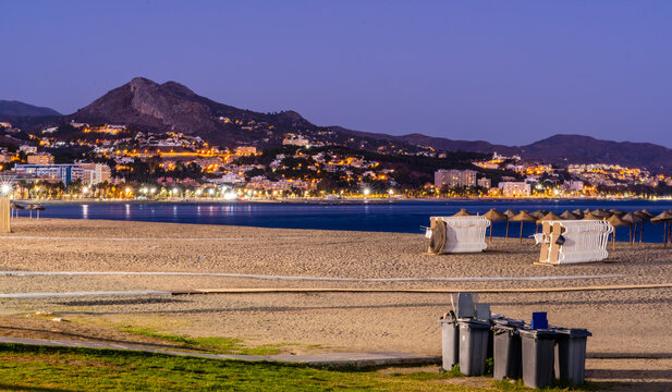 View of the sandy beach at dusk with city lights illuminating the coastline and mountains in the background in Malaga, Andalusia, Spain.