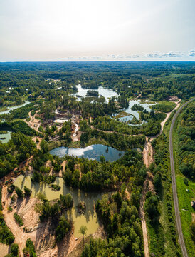 Aerial view of a landscape featuring multiple small ponds, lush green forests, winding dirt paths, and a railway line stretching through the countryside in Latvia.