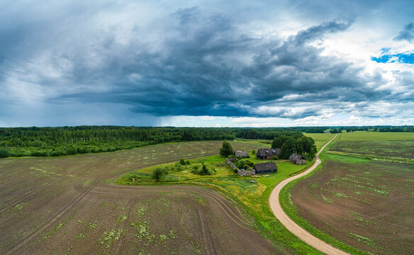 Aerial view of a rural farmstead with traditional wooden buildings surrounded by agricultural fields and a winding dirt road under a dramatic stormy sky in Latvia.