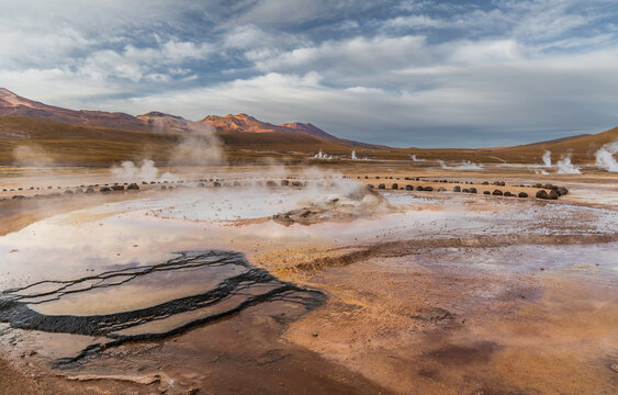 View of El Tatio geyser field with rising steam, geothermal pools, and volcanic mountains under a cloudy sky in Antofagasta, Antofagasta, Chile.