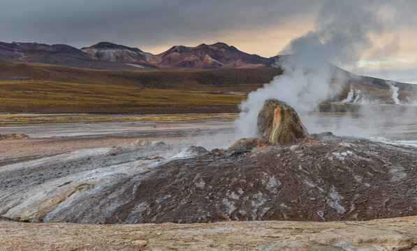 View of El Tatio geyser field with steam rising from volcanic vents against a dramatic mountain landscape in Antofagasta, Antofagasta, Chile.