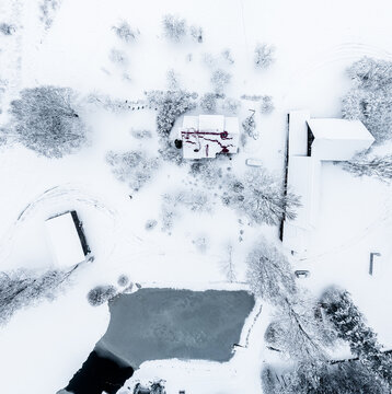 Aerial view of a snow-covered landscape featuring farm buildings and a frozen pond surrounded by trees in Latvia.