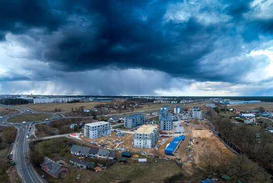 Aerial view of a construction site featuring new apartment buildings, a nearby roundabout, and surrounding rural landscape under a dramatic dark stormy sky in Latvia.