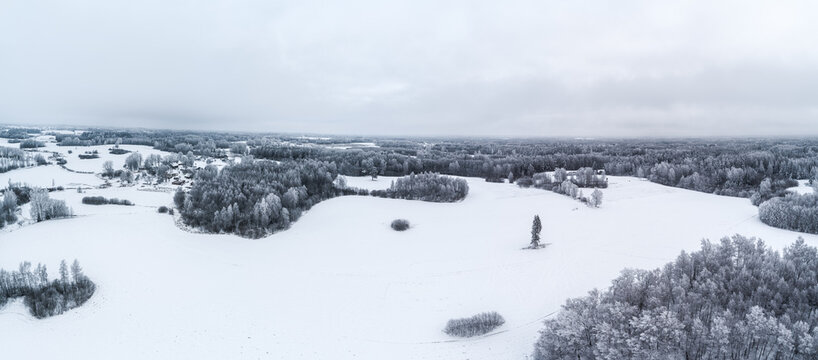 Aerial view of a snow-covered landscape with frozen fields and frost-dusted forests under a cloudy winter sky in Latvia.