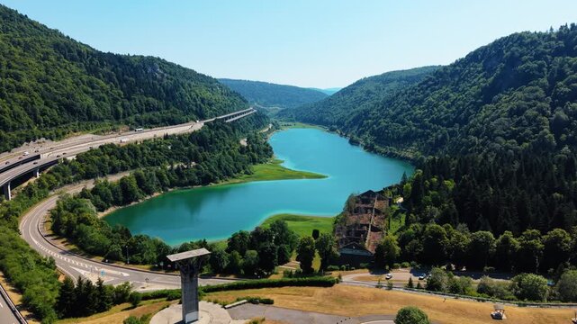 Aerial view of the turquoise Lac de Sylans surrounded by lush green mountains and the A40 highway winding through the landscape in Ain, Auvergne-Rhone-Alpes, France.