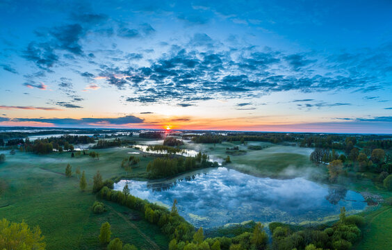 Aerial view of a serene landscape in Latvia featuring a misty lake surrounded by lush green fields and trees at sunrise under a vibrant sky.