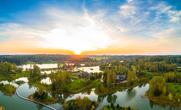 Aerial view of a serene landscape featuring interconnected lakes, lush green forests, and wooden buildings during a vibrant golden hour sunset in Latvia.
