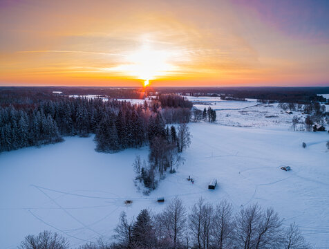 Aerial view of a snow-covered landscape with dense pine forests and open fields under a vibrant sunset sky in Latvia.