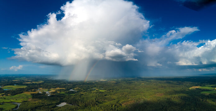 Aerial view of a large cumulonimbus cloud with a rain shower and a faint rainbow over the lush green forests and fields of Latvia.