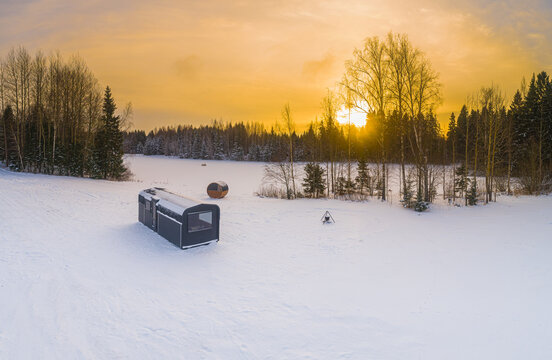 Aerial view of a modern modular cabin and a small round sauna pod in a snow-covered landscape with a fire pit and forest under a golden sunset in Latvia.