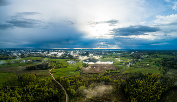 Aerial view of a rural landscape featuring lush green fields, dense forests, a small lake, and a winding dirt road under a dramatic sky with sunbeams breaking through clouds, Latvia.