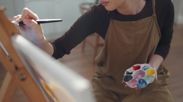 Creative young Asian woman artist painting on canvas with brush in bright home studio. Relaxed female hobbyist enjoying peaceful indoor leisure time, expressing imagination for art.