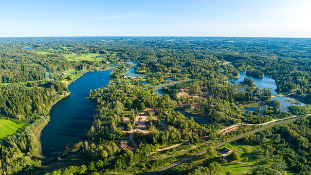 Aerial view of a winding lake surrounded by dense green forests and scattered trees under a clear blue sky in Latvia.