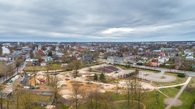 Aerial view of a town featuring a park area under development, residential buildings, roads, and a river under a cloudy sky in Latvia.