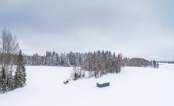 Aerial view of a snow-covered field with a small wooden structure and a dark rectangular building, surrounded by a dense forest of snow-laden pine trees in Latvia.