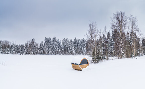 Aerial view of a wooden barrel sauna sitting in a vast snow-covered field with a dense forest of snow-dusted pine trees in the background in Latvia.