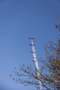 Druskininkai, Lithuania - 23 May 2026: View of four red airplanes flying in a vertical formation with white smoke trails against a clear blue sky seen through tree branches.