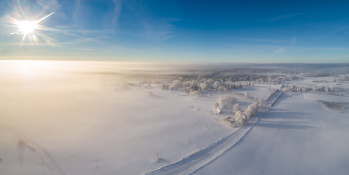 Aerial view of a snow-covered landscape with frost-covered trees and a road emerging from thick morning fog under a bright sun in Latvia.