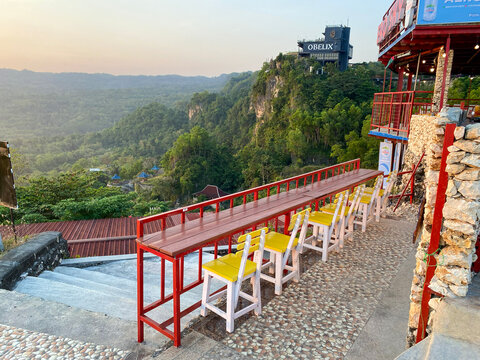 Yogyakarta - Indonesia, &lrm;June 11, 2026: A long wooden bar table with yellow chairs set on a cliffside cafe terrace overlooking a lush green valley and bangunan Obelix.